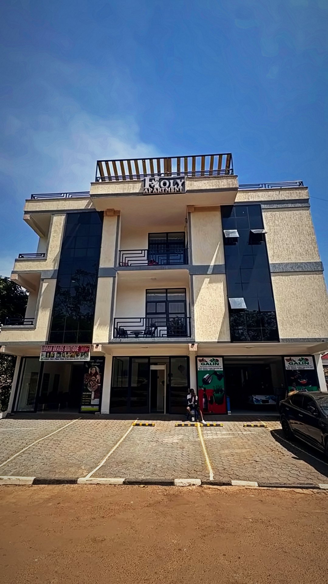 Front of the Holy Apartments building in Kigali with its sign, glass-fronted ground floor and parking