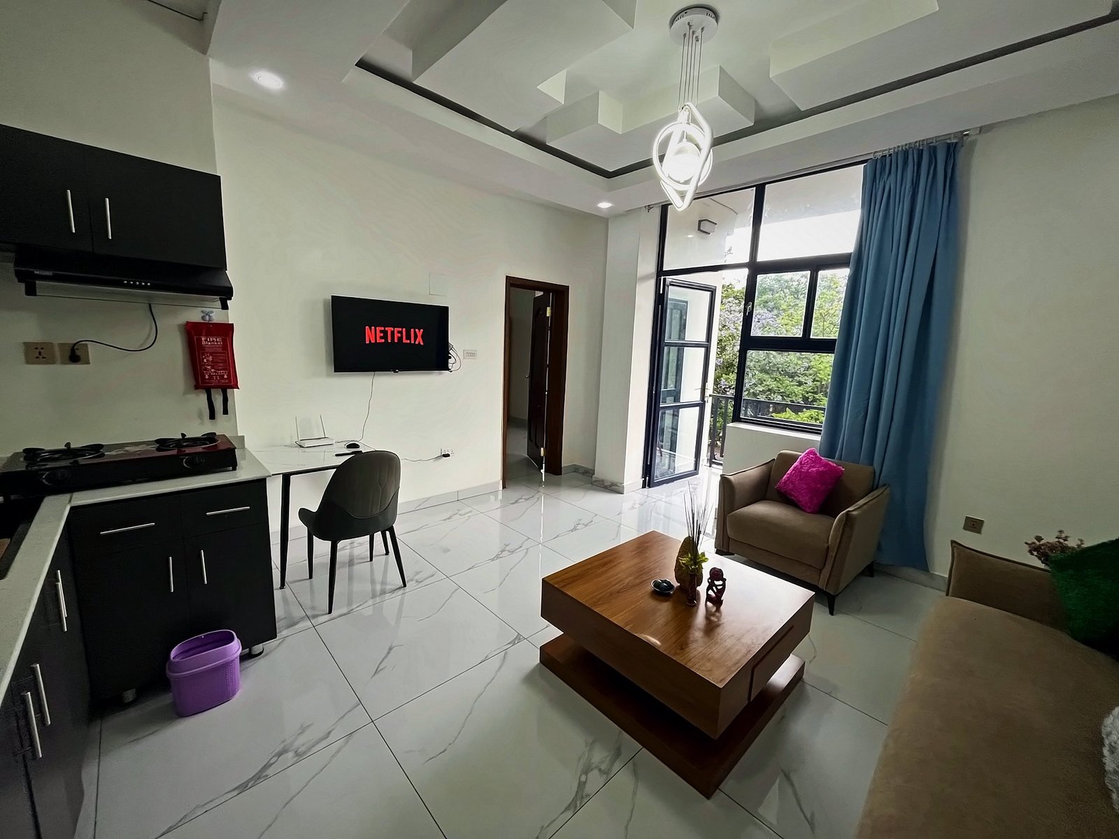 Bright open living area with a sofa, armchair, coffee table, wall-mounted TV and a kitchen corner at Holy Apartments, Kigali
