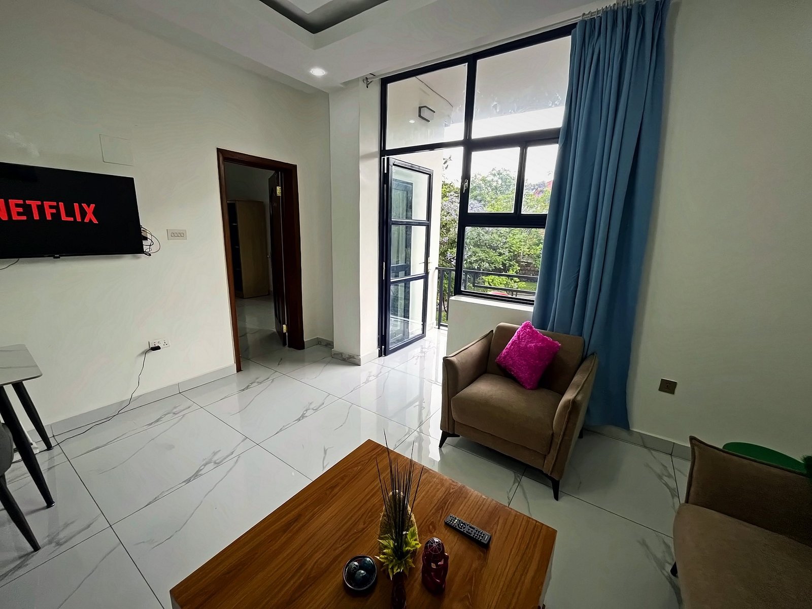 Living room with a wooden coffee table, armchair, TV and a tall window with blue curtains at Holy Apartments, Kigali