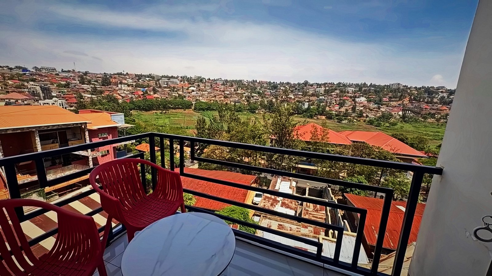 Balcony with red chairs and a table overlooking the red rooftops and green hills of Kigali from Holy Apartments