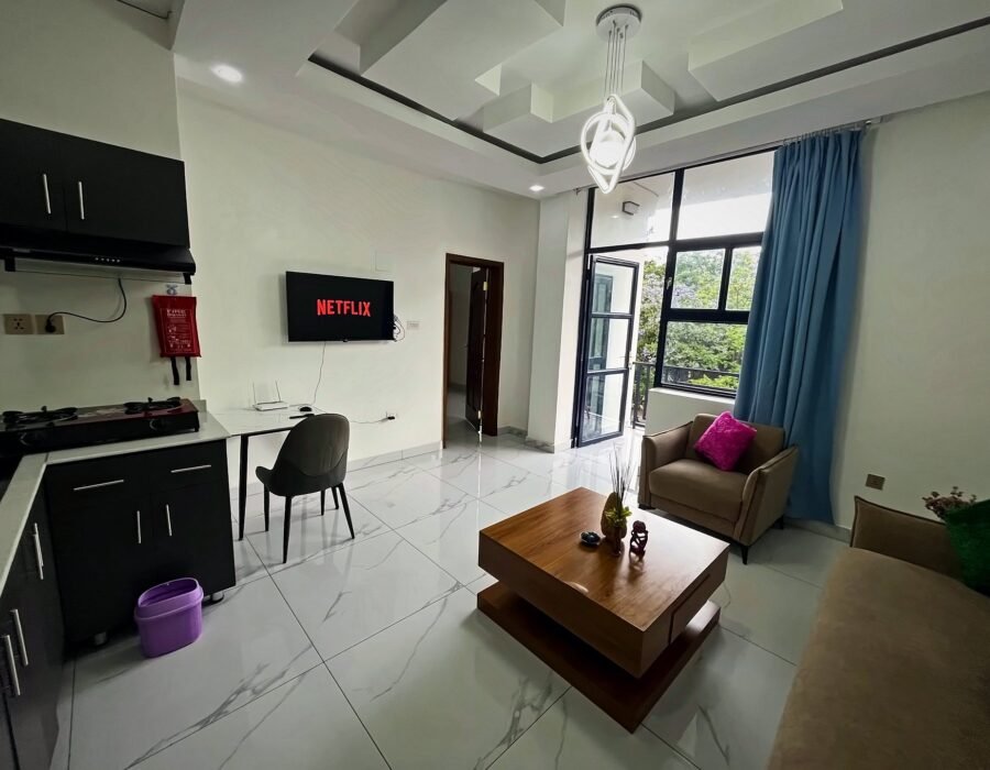 Bright open living area with a sofa, armchair, coffee table, wall-mounted TV and a kitchen corner at Holy Apartments, Kigali