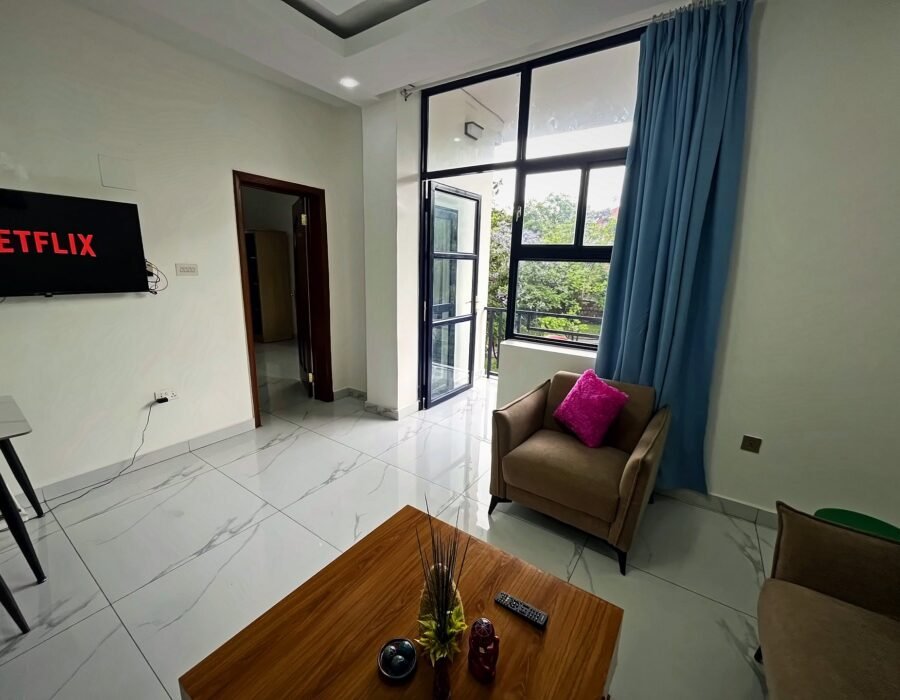 Living room with a wooden coffee table, armchair, TV and a tall window with blue curtains at Holy Apartments, Kigali